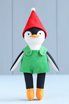 a stuffed penguin with a red hat and green dress is standing in front of a gray wall