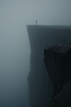 a person standing on top of a cliff in the middle of foggy weather,