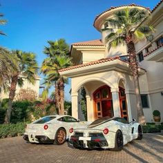 two white sports cars parked in front of a large house with palm trees on the driveway