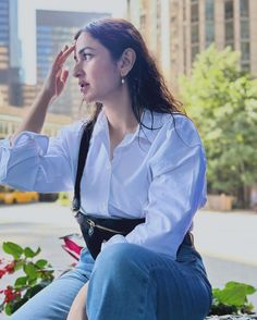 a woman sitting on top of a bench in front of some flowers and buildings with her hand up to her face