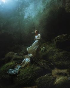 a woman in a white dress is standing on a mossy hill with trees and rocks