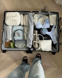 an open suitcase filled with clothing and other items on top of a wooden floor next to someone's feet