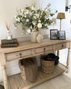 a wooden table with baskets and flowers on it next to a lamp, books and pictures