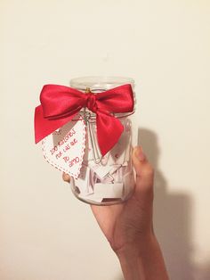 a hand holding a glass jar filled with paper hearts and a red bow on the top