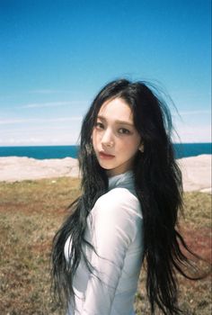 a young woman with long black hair standing in front of the ocean and looking at the camera