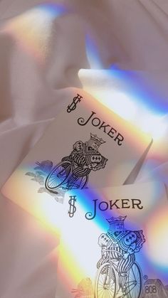 two joker playing cards laying on top of a white sheet with rainbow light coming through them