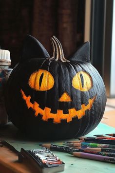 a black cat pumpkin with yellow eyes sitting on a table next to crayons