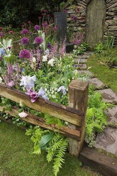 a garden filled with lots of flowers next to a stone wall and wooden bench surrounded by greenery