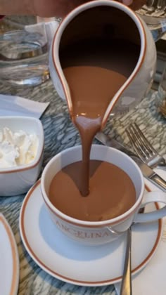 chocolate being poured into a cup with whipped cream on the side and saucer next to it