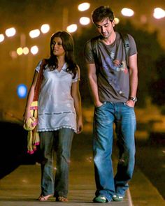 a man and woman walking down a sidewalk at night with lights on the street in the background