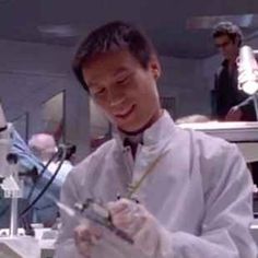 a man in white lab coat and gloves looking at something on a table with other people behind him