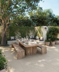 a wooden table sitting under a tree in front of a white wall and some potted plants