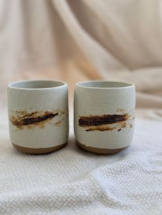 two coffee mugs sitting on top of a bed covered in white sheets and dirt