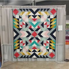 a colorful quilt on display in a room with white walls and curtains behind the wall