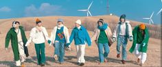 several people are walking in the desert with wind turbines behind them and one person is wearing a green coat