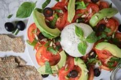 a white plate topped with tomatoes, avocado and black olives next to crackers