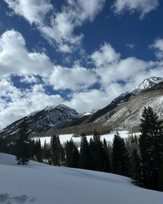 snow covered mountains and trees under a cloudy blue sky with white clouds in the distance