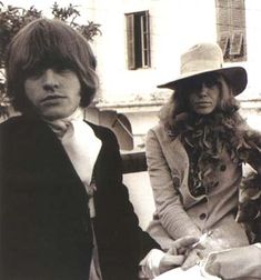 two people standing next to each other in front of a building, one wearing a hat and the other with long hair