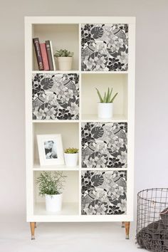 a white bookcase with black and white wallpaper on the front, two planters in