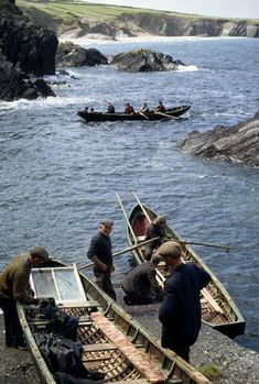 several people in canoes on the shore with rocks and water behind them, one man is pulling something out of the boat