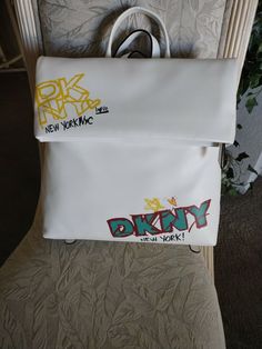 AUTHENTIC “ DKNY” WOMEN’S TILLY WHITE SIGNATURE MD FOLDOVER LEATHER BACKPACK! BRAND NEW/W/TAGS! NEVER USED IN PERFECT CONDITION! STYLE # R12Q350 COLOR: WHITE ONE SIZE M.S.R.P. 168.00 PLUS TAX! FOLDOVER  ZIPPER OPEN/CLOSE W/MAGNETIC SNAP IN CENTER ZIPPER HAS LEATHER PULL TAB JUST UNDER THE FOLDOVER IS A LARGE ZIPPER POCKET THAT  MEASURES: 9” (L) 10.5” (H) ALSO W/WHITE LEATHER PULL TAB SILVER TONE HARDWARE  FRONT OF THE BACKPACK HAS DESIGNER NAME IN YELLOW AND SAYS” LOVE NEW YORK, N Trendy White Bags For Streetwear, Trendy White Streetwear Bags, Trendy White Leather Backpack, Trendy White Backpack, White Backpack For Shopping, Trendy Logo Backpack Bag, Trendy Logo Backpack, Designer Name, R P