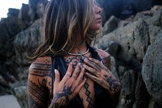 a woman with tattoos on her body standing in front of some rocks and looking off into the distance