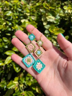 a person is holding three small square earrings in their left hand, and there are green bushes behind them