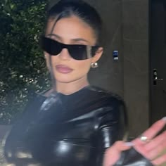 a woman in black leather outfit and sunglasses holding her hand out to the side while walking