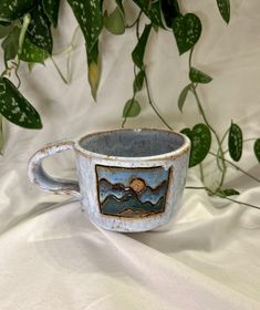 a ceramic cup sitting on top of a white cloth covered table next to a plant