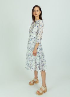 a woman in a white dress and sandals stands against a white background, looking at the camera