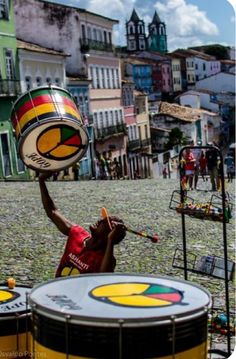 a man is playing drums in the middle of a street with other people and buildings behind him