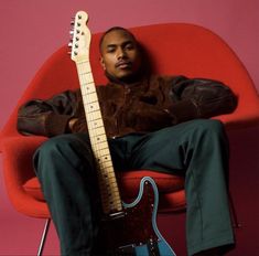 a man sitting in a red chair with a guitar on his lap and looking at the camera