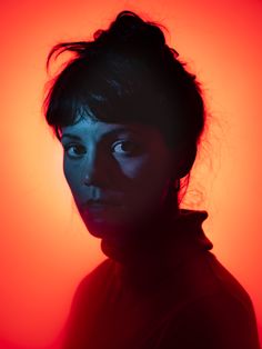 a woman with dark hair and blue eyes in front of a red background is looking at the camera