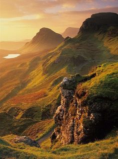 the sun is setting over some mountains with grass and rocks on it, as well as a body of water in the distance