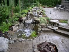 an outdoor fire pit surrounded by rocks and plants with a waterfall in the middle on a brick patio