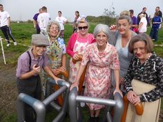 a group of older women standing next to each other on a dirt road with people in the background