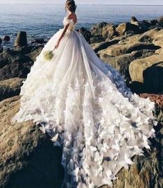 a woman is standing on some rocks by the ocean wearing a wedding dress with feathers