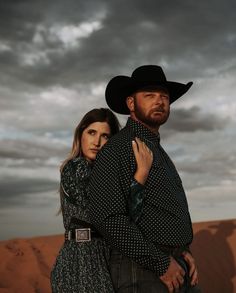 a man and woman standing in the desert under a cloudy sky with their arms around each other