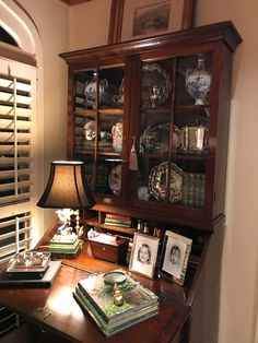 a wooden desk topped with lots of books next to a lamp and window covered in shutters