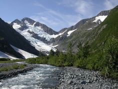 a river running through a lush green forest filled with snow covered mountains in the distance