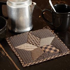 a black table with a coffee pot and two mugs on it, next to a quilted coaster