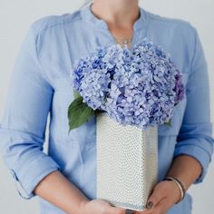 a woman holding a white vase with blue flowers in it's center, while wearing a blue shirt