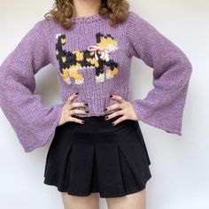 a woman wearing a purple sweater and black skirt with her hands on her hips, standing in front of a white wall