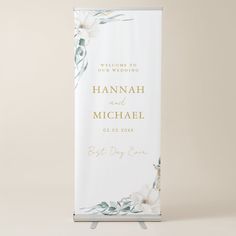 a white and gold wedding welcome sign with flowers on the front is shown against a neutral background