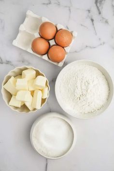 eggs, butter and flour in bowls on a white marble countertop next to an egg carton