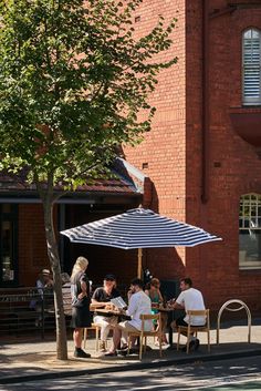 people are sitting at tables under an umbrella on the side of the road in front of a brick building