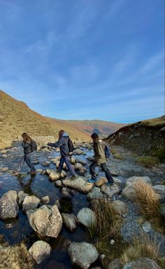 three people cross a stream on rocks in the middle of a grassy area with mountains in the background