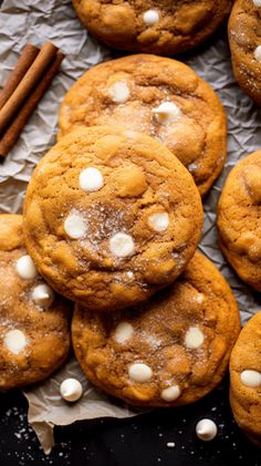 a pile of cookies with white chocolate chips and cinnamon sticks next to it on a piece of parchment paper