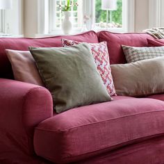 a red couch with several pillows on it in front of two windows and a lamp