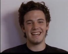 a man with curly hair smiling and wearing a black t - shirt in front of a white wall